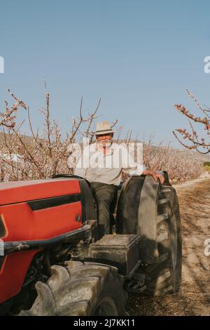 Senior male farmer sitting in tractor while working in agricultural plantation with blooming apricot trees in countryside on summer day looking at cam Stock Photo