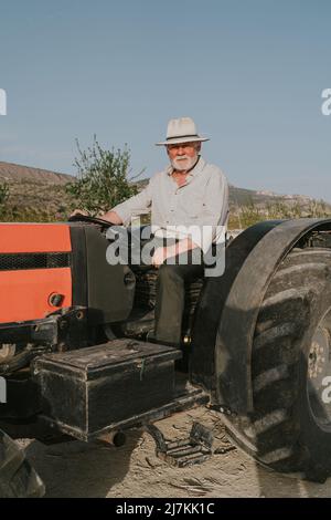 Senior male farmer sitting in tractor while working in agricultural plantation with blooming apricot trees in countryside on summer day looking at cam Stock Photo