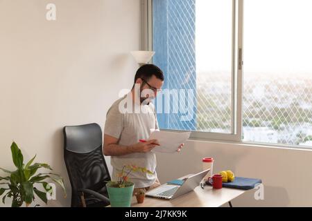 Focused bearded male worker in eyeglasses and casual wear reading documents at table with laptop while working in light home office Stock Photo