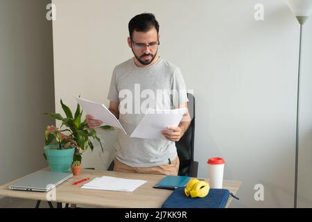 Focused bearded male worker in eyeglasses and casual wear reading documents at table with laptop while working in light home office Stock Photo