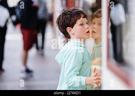 Side view of curious little boy in turquoise hoodie with dark hair leaning on glass door and trying to peek inside train while standing on platform of Stock Photo