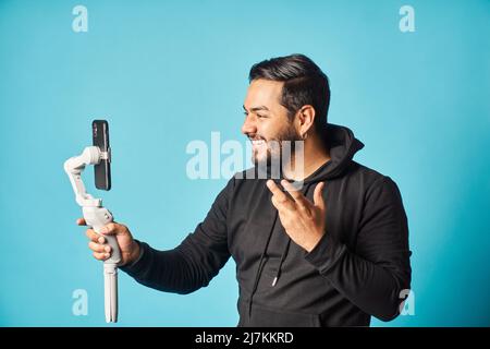 Positive bearded male blogger in black hoodie recording vlog on modern smartphone while standing against blue background in light studio Stock Photo