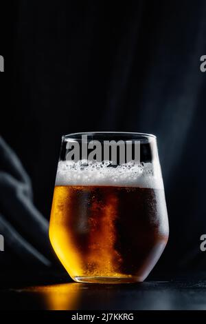 Transparent glass filled with brown alcoholic beer and white froth served on dark table in light room against black background