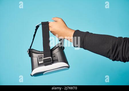 Hand of crop anonymous gamer holding modern VR headset for exploring cyberspace while standing on blue background in light studio Stock Photo