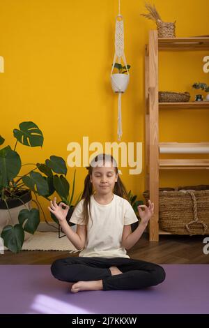 focused young girl in activewear sitting on mat in Padmasana and meditating while practicing yoga at home Stock Photo