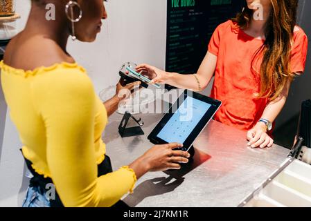 Crop young female customer scanning smartphone for paying while standing at cafe counter with African American barista checking bill on tablet