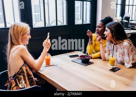 Happy young blonde Woman taking picture with smartphone of diverse female friends eating smoothie while sitting together at table in cafe and having b Stock Photo