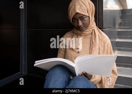 Crop concentrated young African American Muslim female student in traditional headscarf sitting on street and reading book while preparing for exams Stock Photo