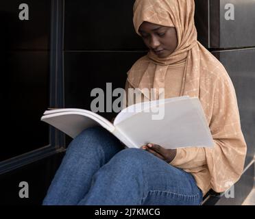 Crop concentrated young African American Muslim female student in traditional headscarf sitting on street and reading book while preparing for exams Stock Photo