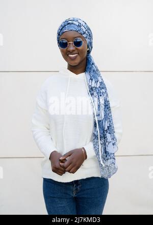 Self assured young African American female millennial in trendy sunglasses and traditional headscarf smiling and looking at camera against white wall Stock Photo