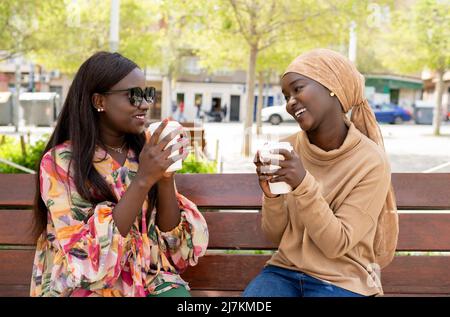 Positive young African American female best friends in casual outfits smiling and looking at each other while drinking takeaway coffee sitting on benc Stock Photo