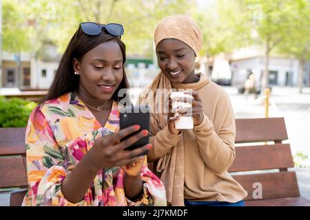 Stylish positive young African American Woman with long dark hair using mobile phone while sitting on bench with smiling Muslim friend drinking takeaw Stock Photo