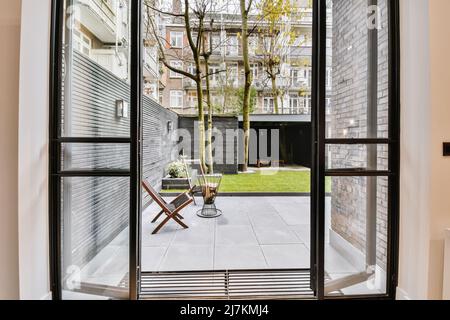 Opened glass doors of room overlooking terrace with chairs and lawn against residential building located in town on summer day Stock Photo