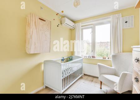 White crib with comfortable big armchair placed near window with toys on windowsill in light spacious children bedroom at home Stock Photo