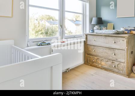 White crib placed at wall near window in light spacious children bedroom with wooden chest of drawers with lamp at home Stock Photo