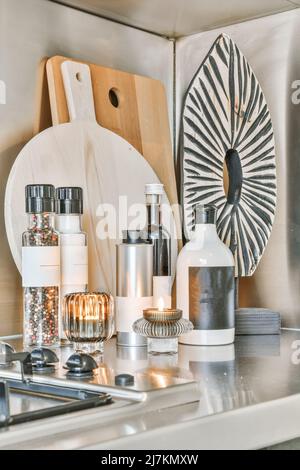 Bottles with assorted supplies and burning candles placed near wooden cutting boards and cooker on counter in light modern kitchen Stock Photo