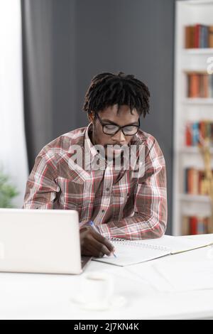 Young african student man making notes working at home working on laptop or listening teacher or boss during video call. African student study using laptop at home. Stock Photo