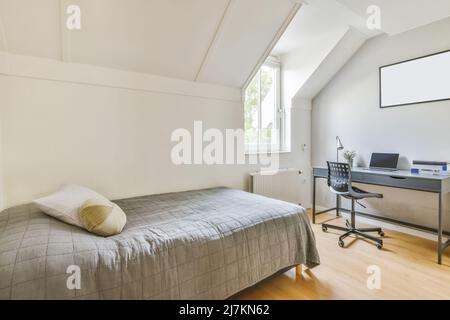 Comfortable bed with gray blanket placed near table with netbook and lamp at glass window in light bedroom of apartment Stock Photo