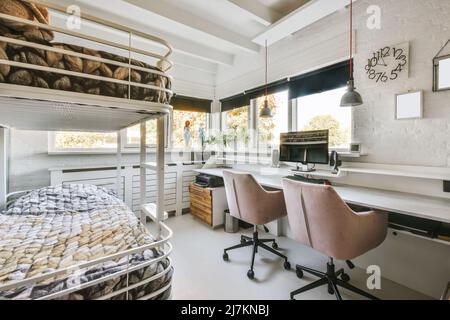 White bunk bed with soft mattresses in spacious teen room with chairs at working table with computer placed near window in daylight in modern apartmen Stock Photo