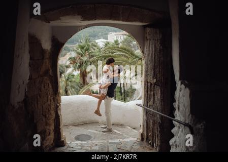 View through old stone arch of loving man carrying Woman in dress while standing on step in tropical town Stock Photo