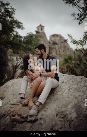 Tender casual couple cuddling while sitting on rock at green mountain valley with distant building Stock Photo