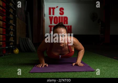 Concentrated young athletic Woman with dark hair in sportswear doing plank exercise on mat and looking at camera during workout in modern gym Stock Photo