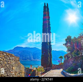 The hilly street of Morcote with a view of the old stone wall, tall cypress trees, Lake Lugano and Alps in the background, Switzerland Stock Photo