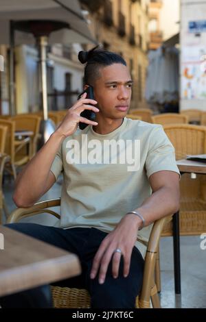 Serious young Hispanic male in casual clothes having phone conversation while sitting on terrace of outdoor cafeteria on summer day Stock Photo