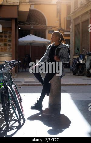 Full body side view of young Hispanic male sitting on pillar near bicycle parked on sidewalk on street with residential buildings Stock Photo
