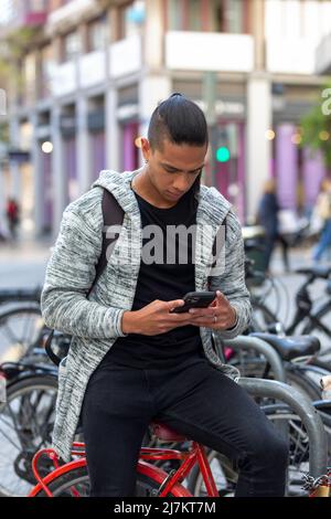 Focused Hispanic male in casual wear texting message on cellphone while sitting on bicycle on street of city with building Stock Photo