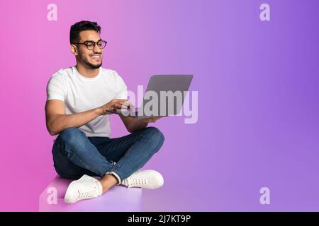 Full length of millennial Arab man in glasses using laptop for work or studies in neon light, copy space Stock Photo
