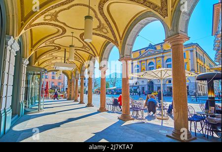 LUGANO, SWITZERLAND - MARCH 25, 2022: The scenic arcade of historic building on Piazza della Riforma square opens the view on the square, outdoor cafe Stock Photo