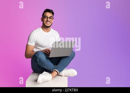 Young Arab man in casual wear working online, sitting cross legged and using laptop in neon light, full length Stock Photo