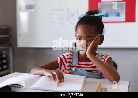 Sad african american girl with hand on cheek and books sitting at desk in classroom. unaltered, education, learning, studying and school concept. Stock Photo