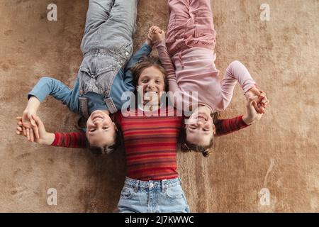 Top view of happy three sisters kids lying on floor and looking at camera. Stock Photo