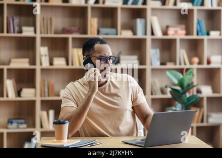 Remote business concept. Concerned arab man talking on cellphone and using laptop while sitting at desk at home Stock Photo
