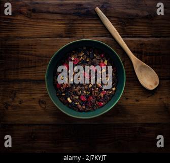 Flat lay composition on a dark wooden table of mixed dried fruit in a dark green bowl, ready to make a fruit cake. Stock Photo