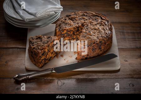 Fruit cake served on a marble board with a segment cut out, on a dark wood work top, with a stack of white plates and napkins in the background. Stock Photo