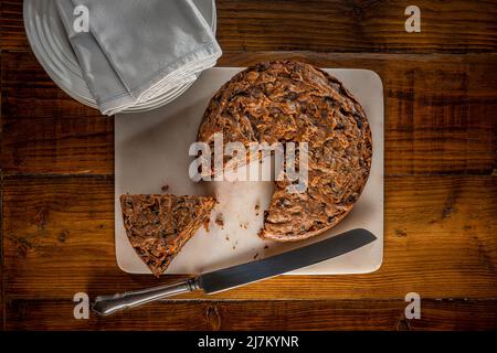 Fruit cake served on a marble board with a segment cut out, on a dark wooden work top, with a stack of white plates and napkins in the background. Stock Photo