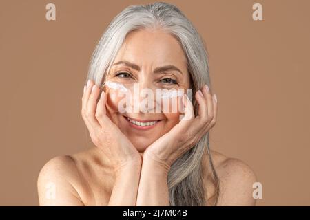 Mature beauty. Charming senior woman posing with face cream under her eyes, smiling at camera over brown background Stock Photo