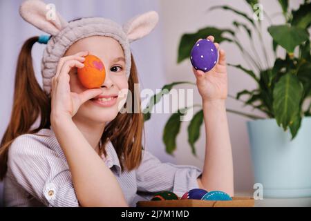 A positive playful girl in bunny ears holds colorful eggs in her hands in the home kitchen. Happy Easter. Easter background Stock Photo
