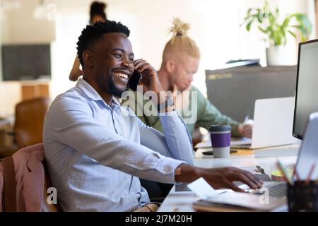Cheerful african american mid adult businessman talking on smart phone while using laptop in office Stock Photo