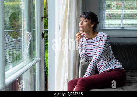 Thoughtful caucasian young woman looking through window while sitting at home Stock Photo