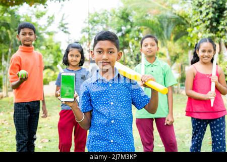 Smiling kid showing green screen mobile phone with cricket bat by looking at camera with friends at park - concept of training, advertisement and Stock Photo