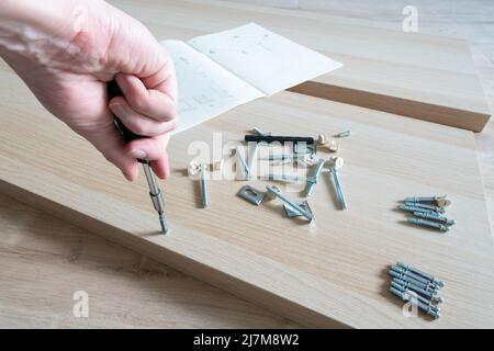 Male right hand and parts of unfinished furniture , metal screws and tools lying on the floor with instruction manual for assembly in the background Stock Photo