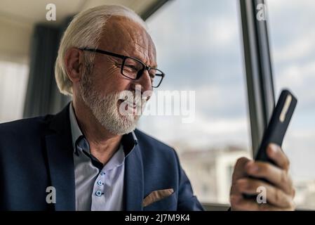 Smiling businessman professional text messaging on mobile phone. Elderly businessman sitting by window working in corporate office. Stock Photo
