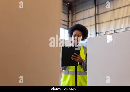 African american young female worker holding clipboard while checking cardboard boxes in warehouse Stock Photo