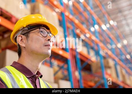 Low angle view of thoughtful asian mature male worker wearing hardhat and eyeglasses looking away Stock Photo