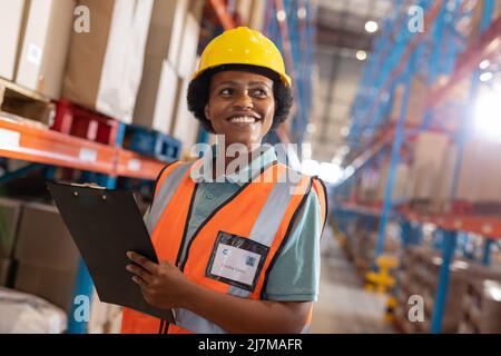 Smiling african american young female worker holding clipboard and looking away in warehouse Stock Photo