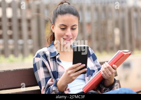 Happy student checking smartphone holding folders sitting in a bench Stock Photo